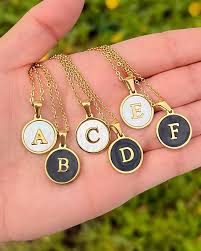 Hand holding five gold necklaces with lettered pendants against a blurred green background