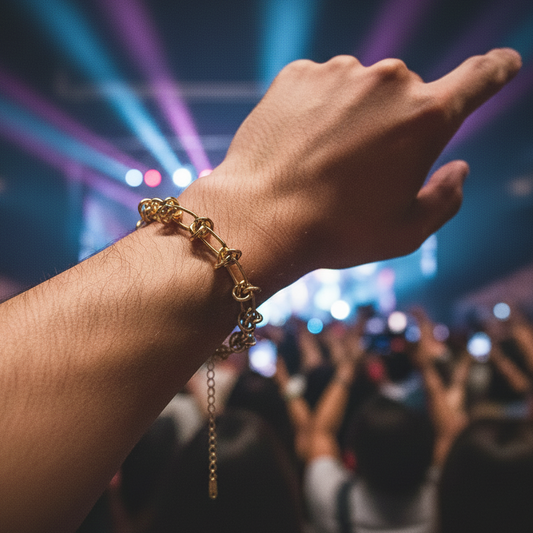 A close-up of a person's wrist wearing a gold chain bracelet with a small heart charm, pointing toward a concert stage with colorful stage lights in the background.