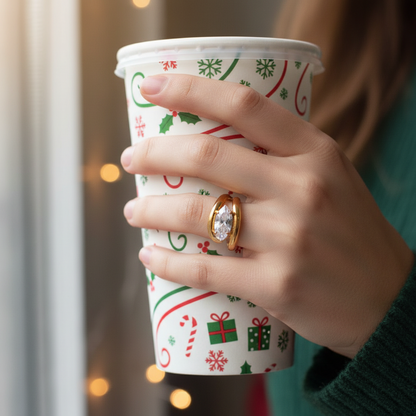 Hand with gold open horizontal solitaire marquis holding a Christmas-themed paper cup with a blurred background