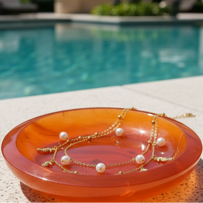A gold-tone double-strand necklace with white pearl beads and small dangling charms, displayed on a translucent orange glass dish against a dark reflective background.