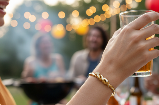 A close-up lifestyle shot of a woman at an outdoor barbecue holding a glass of beer, wearing a gold twisted nail rhinestone bracelet on her wrist. The background shows a blurred outdoor party scene with people, a grill, and warm string lights.