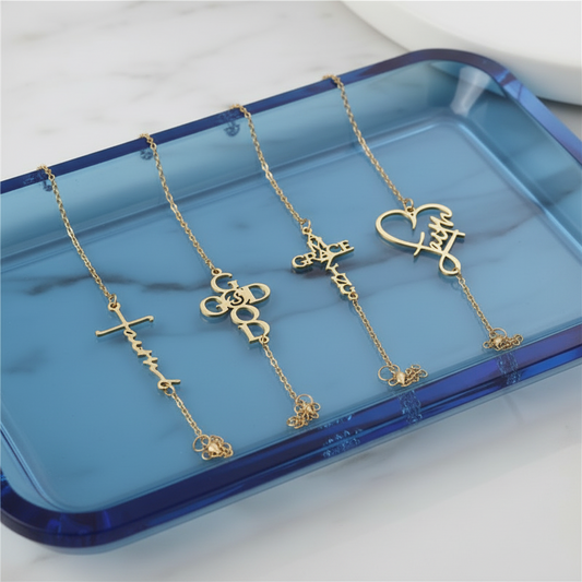 Four gold-tone nameplate necklaces with different script pendant designs, displayed on a black glittery tray.
