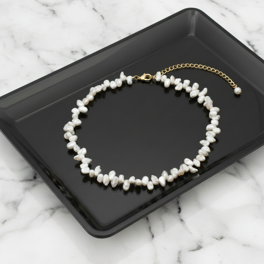 A necklace made of irregular white freshwater pearls with a gold chain extender and clasp, displayed on a glossy black rectangular tray against a white background.