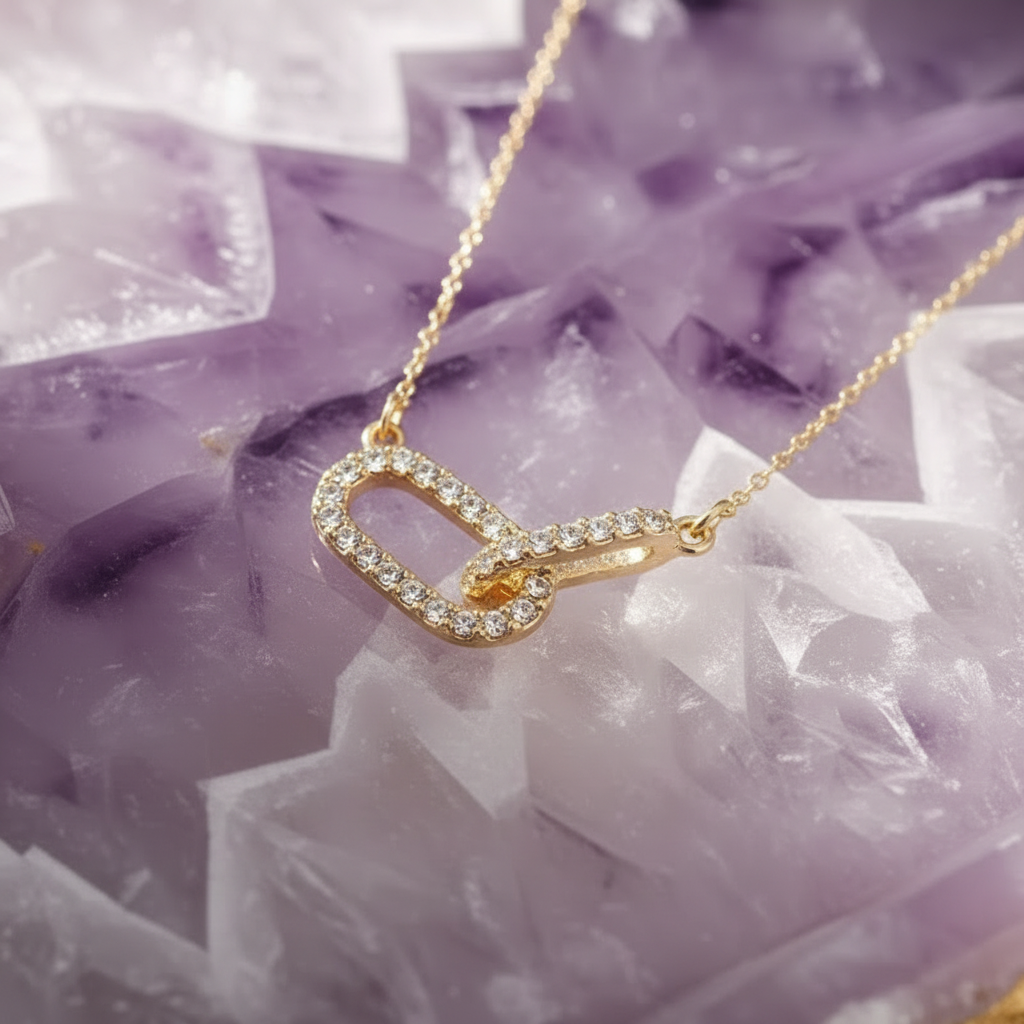 A delicate gold necklace with a small, openwork pendant encrusted with clear stones and a tiny orange accent, displayed on a purple amethyst geode slice.