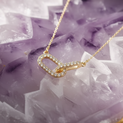 A delicate gold necklace with a small, openwork pendant encrusted with clear stones and a tiny orange accent, displayed on a purple amethyst geode slice.